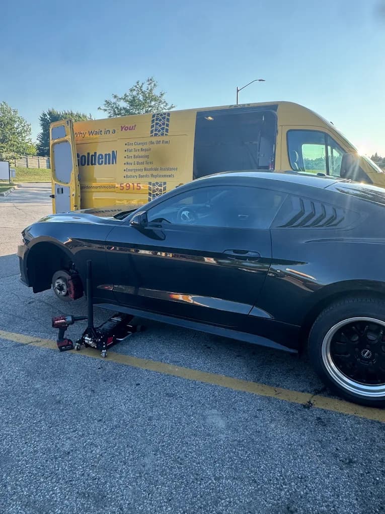 Black Mustang on the jack for a tire change, yellow van behind on a sunny day