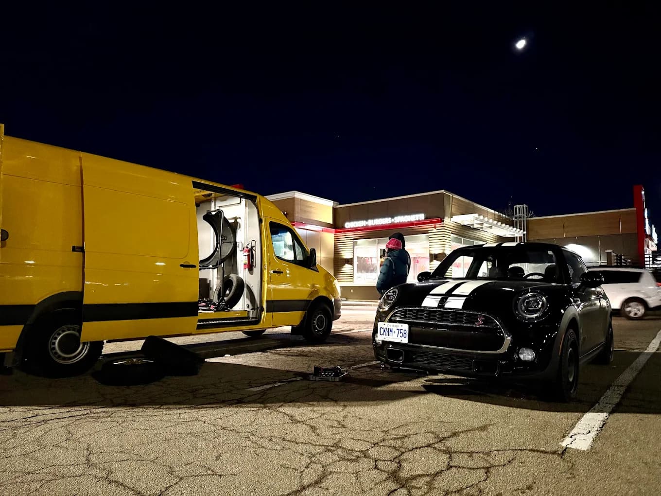 Night mobile battery and jump start service for a MINI at a lit gas station, the open yellow van alongside